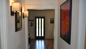 A hallway with a wooden floor, a door with vertical blinds, wall-mounted lamps, and a framed picture on the right wall. - Very clean house with amazing park across the street and Japanese garden. (Coos Bay)