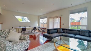 A spacious living room with a large black leather sofa, a patterned armchair, and a glass coffee table. There is a dining area with a table and chairs, and large windows providing natural light. - Very clean house with amazing park across the street and Japanese garden. (Coos Bay)