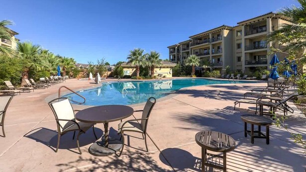 A pool area with lounge chairs and tables, surrounded by palm trees and a building with balconies.