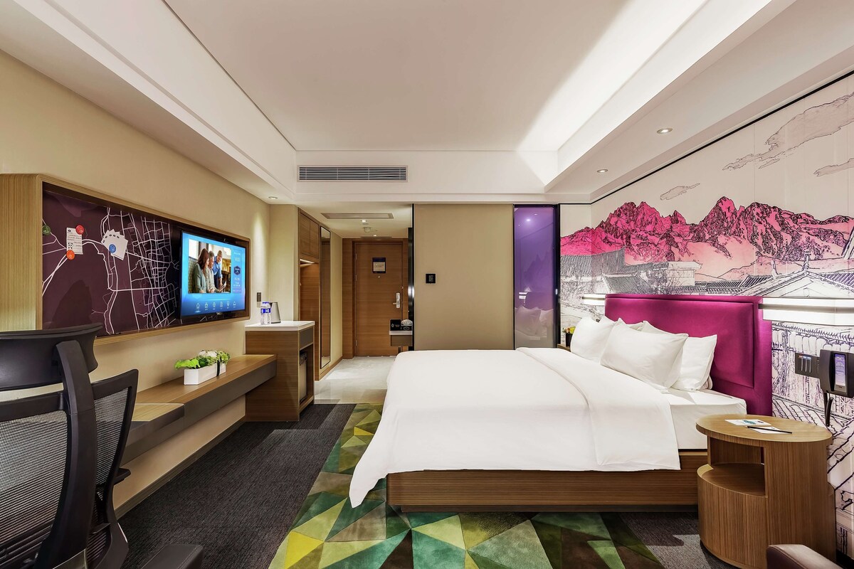 A modern hotel room with a bed, desk, chair, and a TV on the wall.
