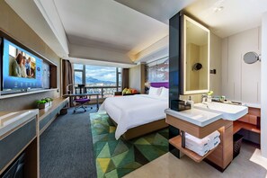A hotel room with a large bed, a desk with a computer, a TV, a bathroom with a mirror and sink, and a view of the city. - Hampton by Hilton Lijiang Old Town (Lijiang)