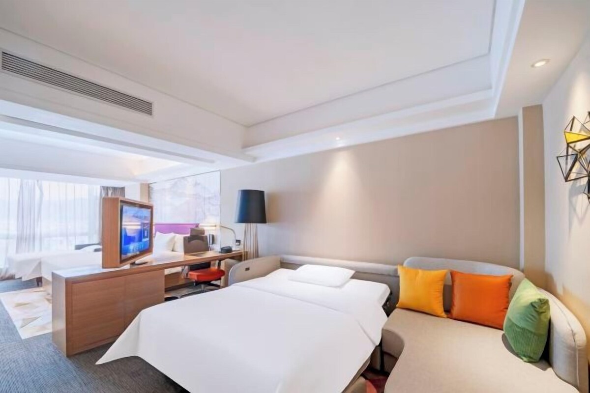A hotel room with a bed, a desk, a TV, a sofa, and a wall-mounted lamp.