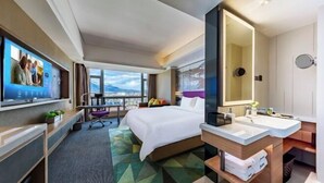 Ein modernes Hotelzimmer mit einem großen Bett, einem Schreibtisch mit Computer, einem Fernseher und einem Badezimmer mit großem Spiegel.