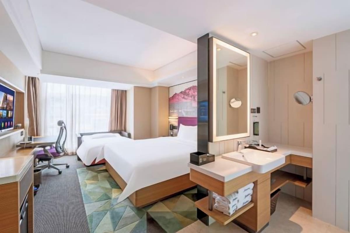 A hotel room with a bed, a desk, a chair, a large mirror, a sink, and a window.
