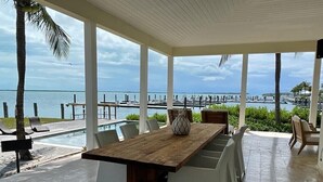 Un espace extérieur abrité, aménagé pour les repas, avec une table en bois et des chaises blanches, offrant une vue sur une marina et la mer.