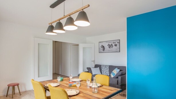 A dining area with a wooden table, yellow chairs, and a blue accent wall. - Appartement Ribera - Welkeys (Annecy)