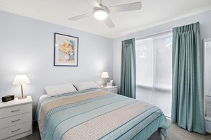 Un dormitorio con cama, dos mesitas de noche con lámparas, ventilador de techo, ventana con cortinas e imagen enmarcada en la pared.