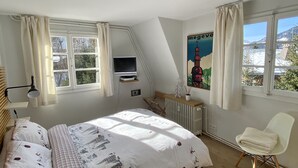 A bedroom with a bed, a chair, a TV, a radiator, and a window with a view of trees and mountains. - Appartement Odyssée - Welkeys (Megève)