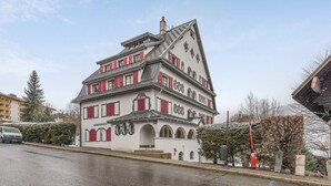 A two-story building with a steeply pitched roof, multiple windows with shutters, and a prominent entrance with an archway. - Appartement Odyssée - Welkeys (Megève)