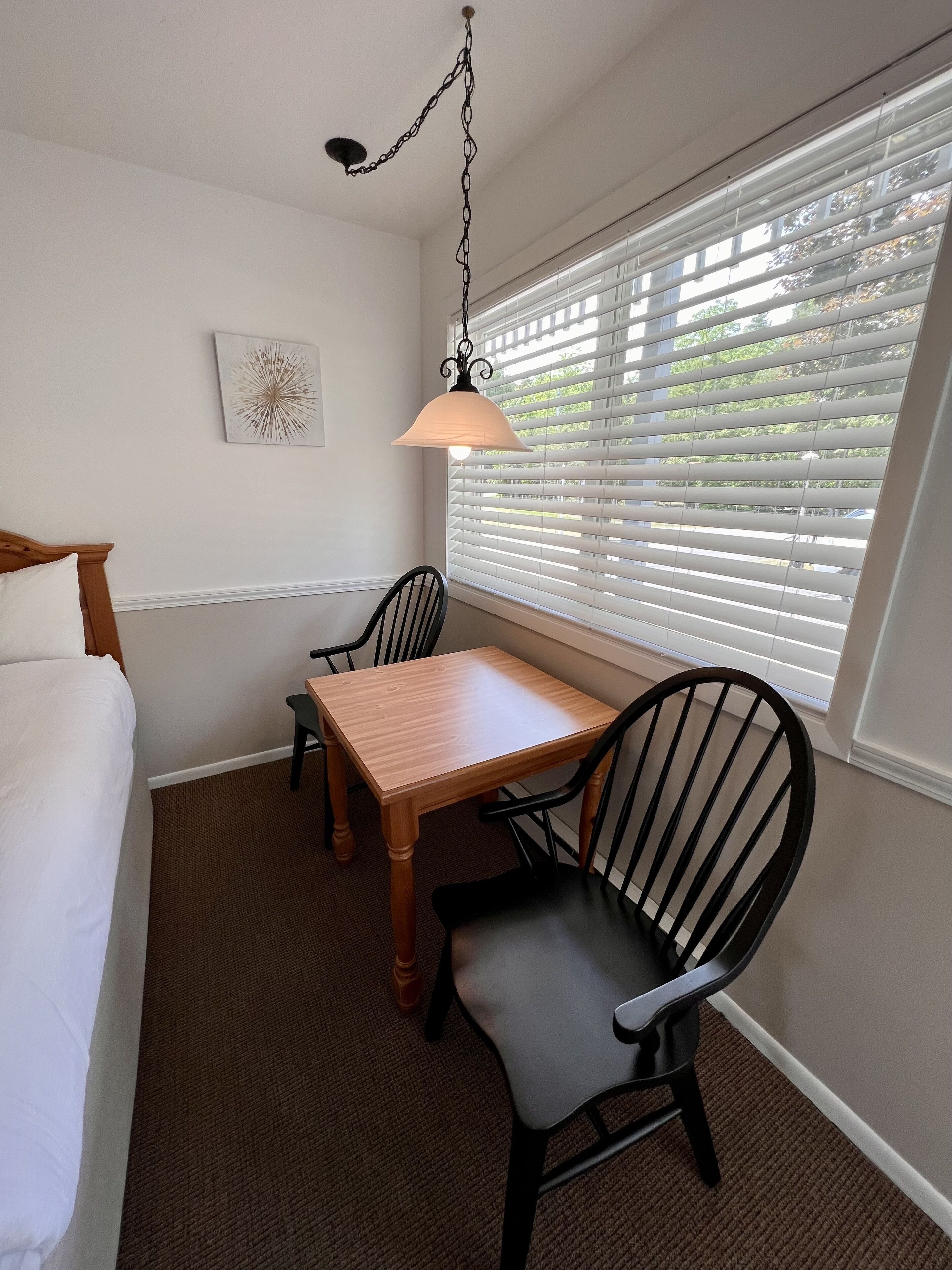 A hotel room with a bed, a wooden table, two chairs, a hanging lamp, and a window with blinds.
