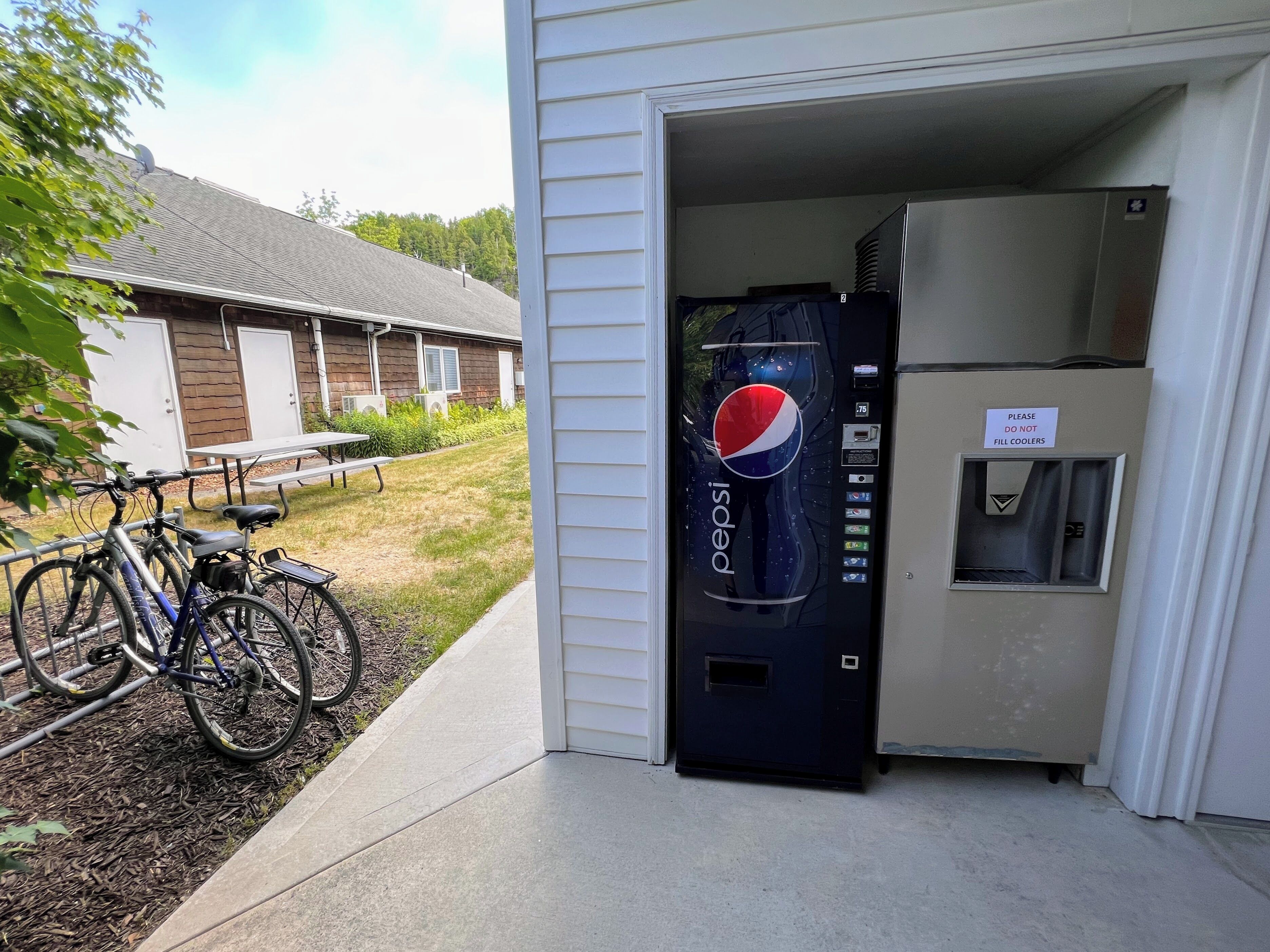 Ein Pepsi-Automaten vor einem Gebäude, in der Nähe stehen ein Tisch und Fahrräder.
