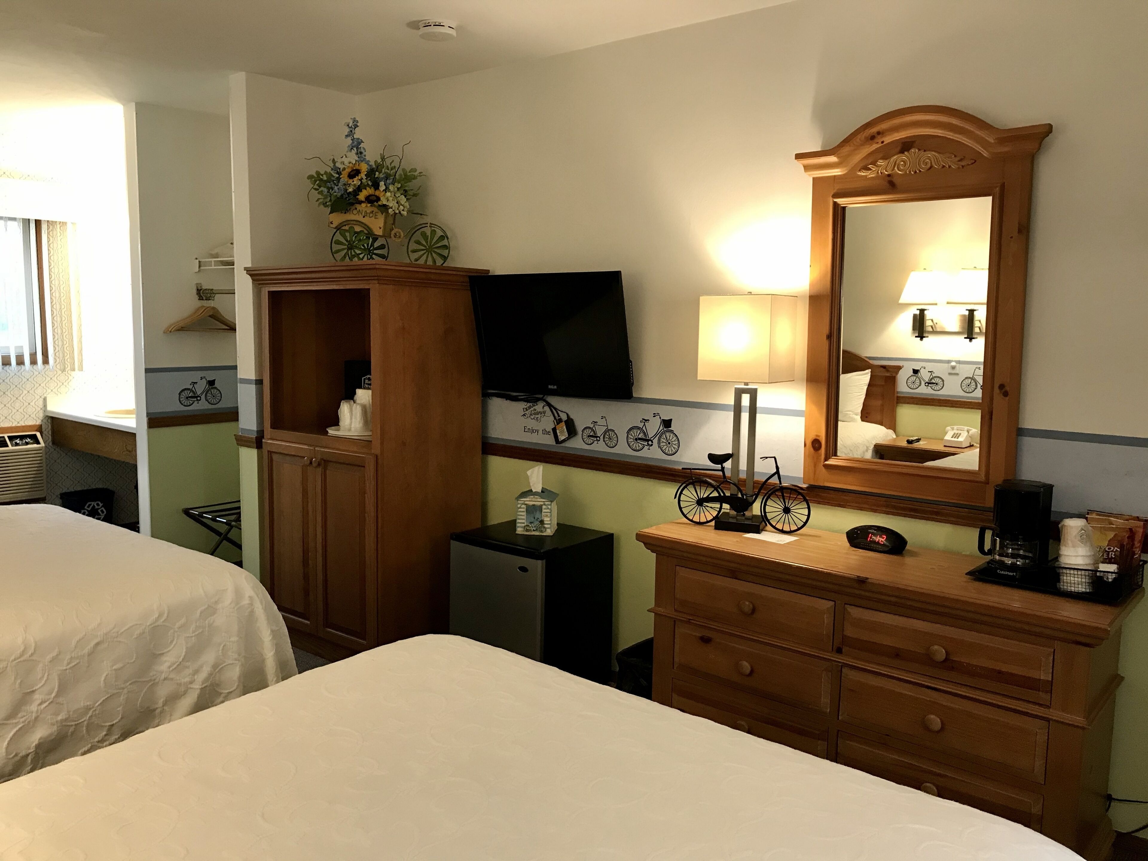 Camera d'albergo con due letti, una cassettiera in legno, uno specchio, un televisore e una decorazione a forma di bicicletta appesa al muro.