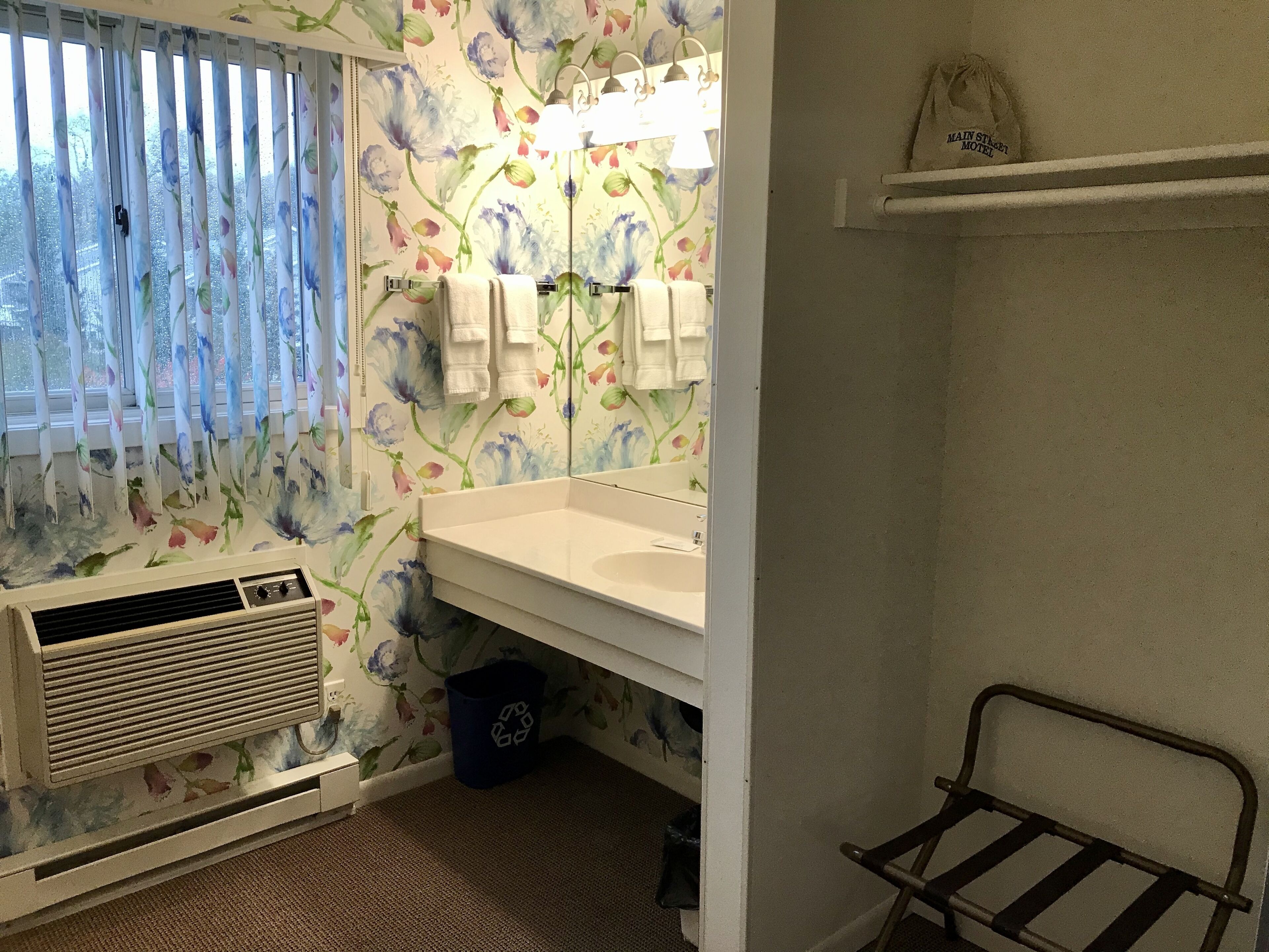 Een badkamer met bloemenbehang, een witte wastafel, een spiegel, een handdoekhouder, een verwarming, een afvalbak voor recycling en een metalen rek.
