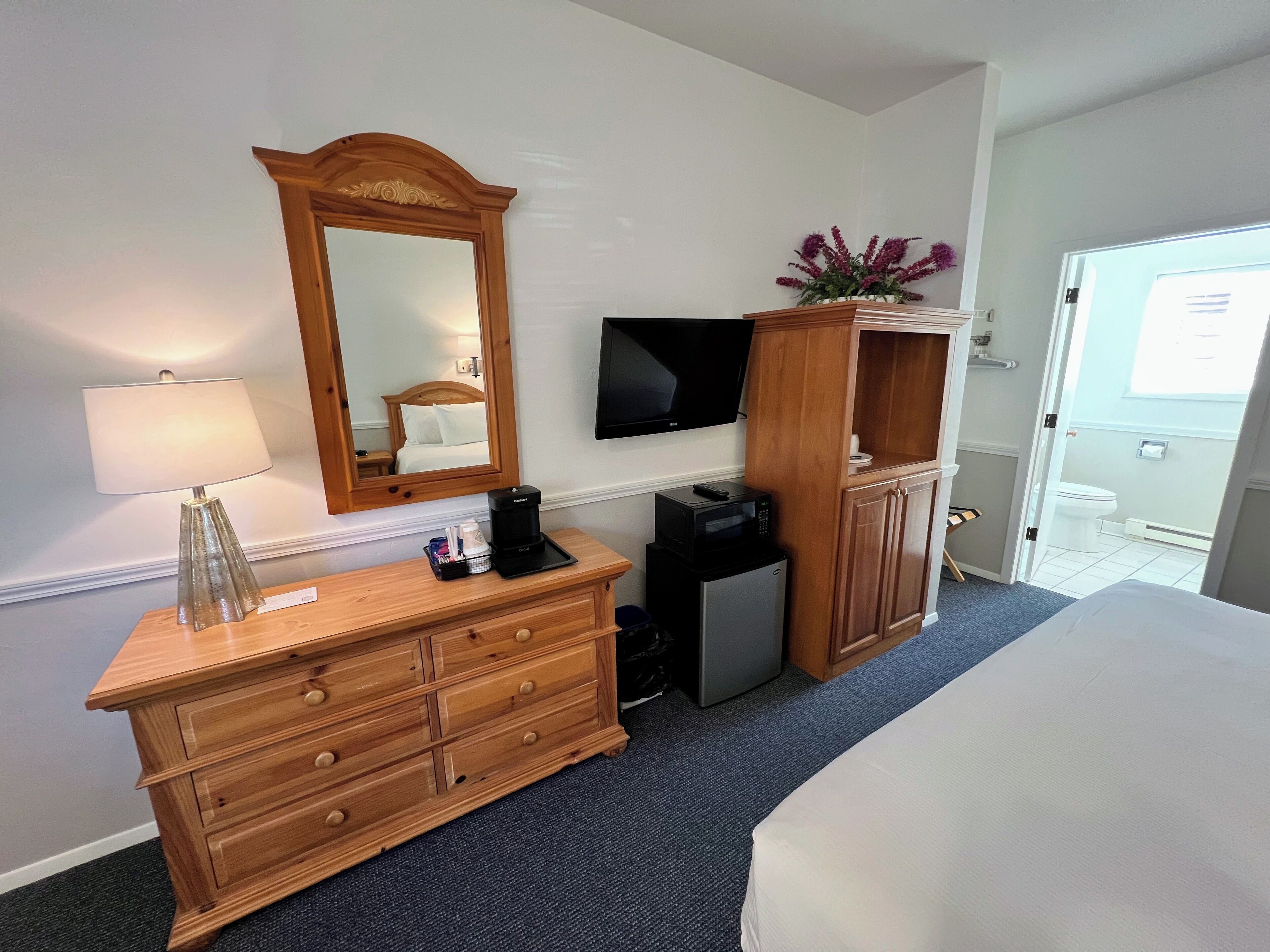 A hotel room with a wooden dresser, a mirror, a flat-screen TV, a coffee maker, a microwave, and a small cabinet with a flower arrangement.