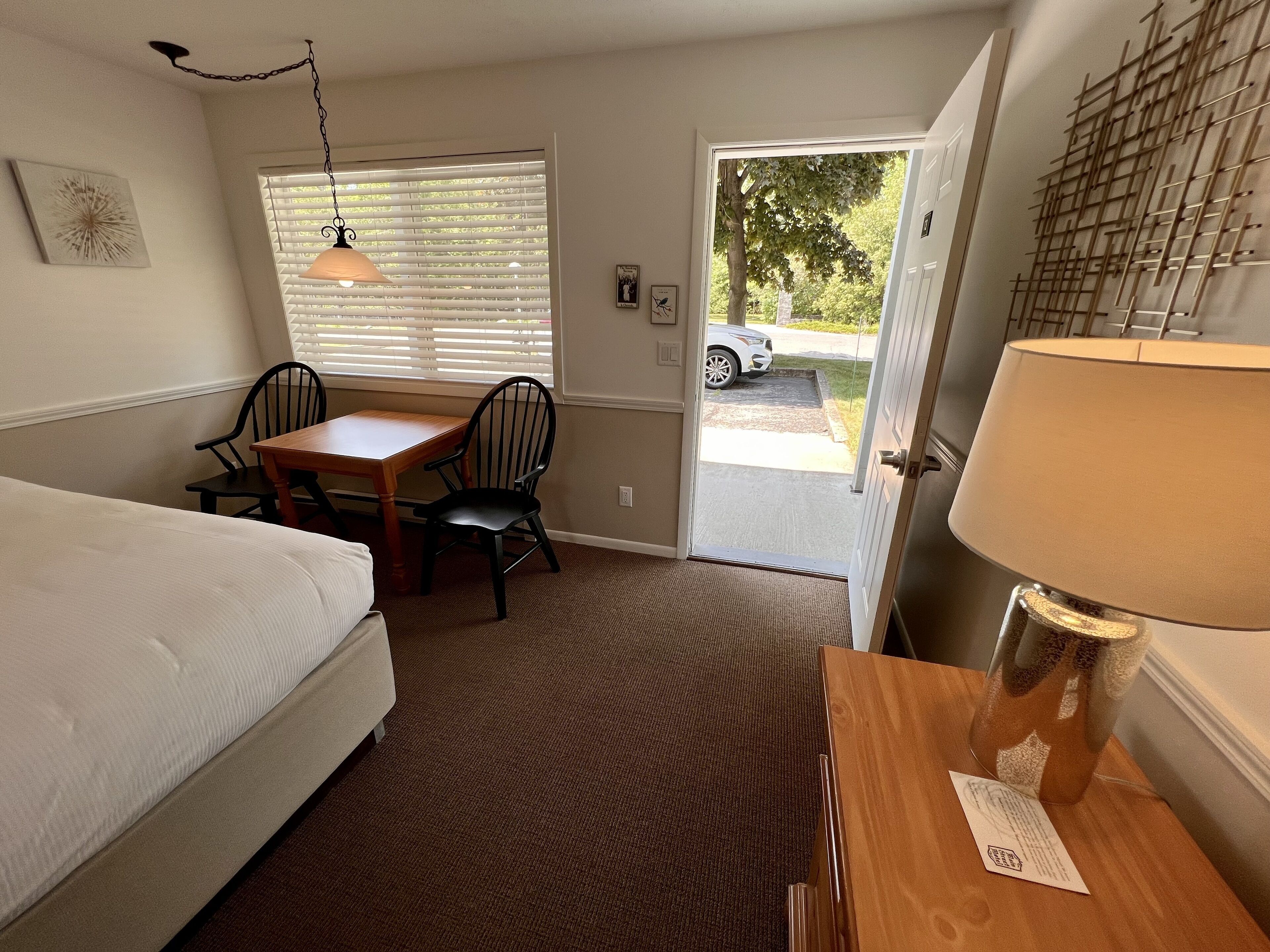 Habitación de hotel con cama, una mesa de madera, una silla, una lámpara, una ventana con persianas y una puerta que da al exterior.