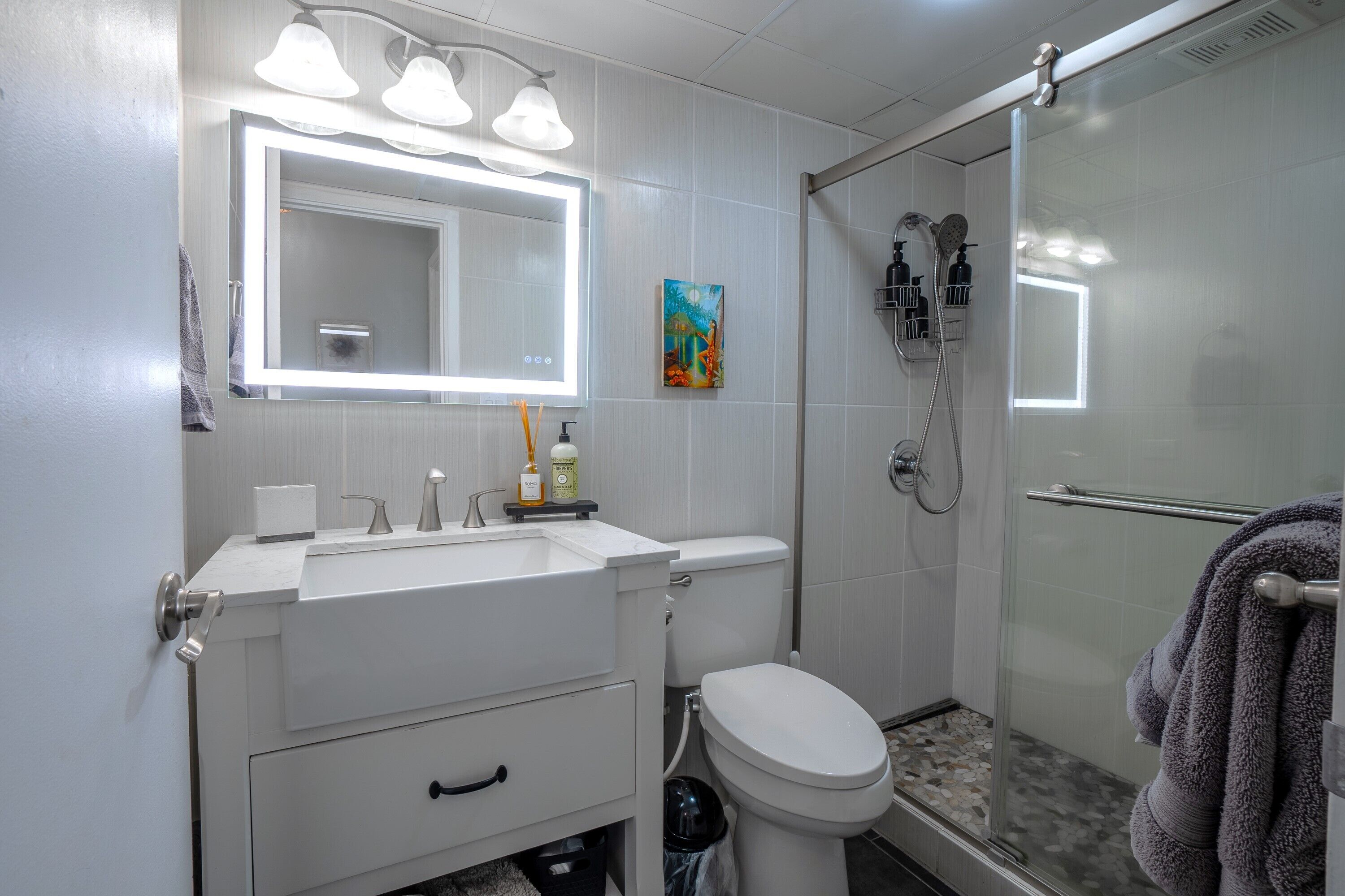 Une salle de bain moderne avec un lavabo, un miroir et des toilettes blancs. Elle comprend un espace douche avec une porte en verre et un porte-serviettes.