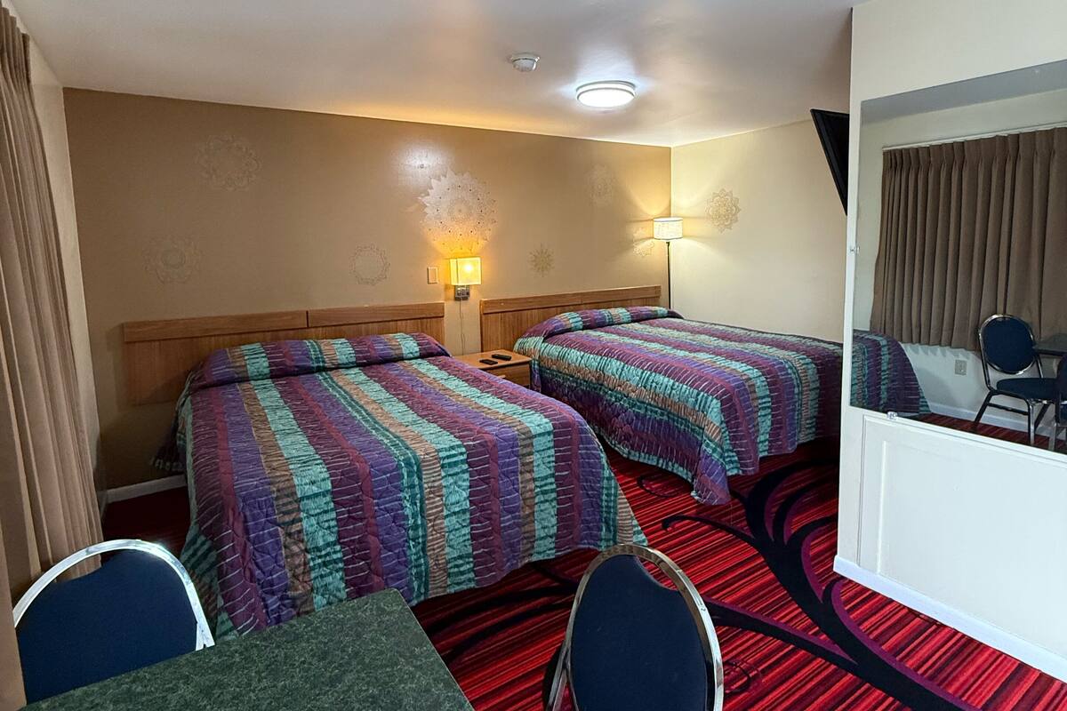 Habitación de hotel con dos camas, una mesita y sillas. Se observa un espejo en la pared y una lámpara en el techo.