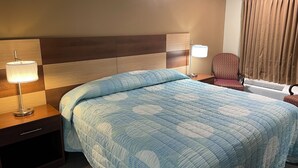 Habitación de hotel con una cama grande, una mesita de noche con lámpara, una silla y un ventanal con cortinas.