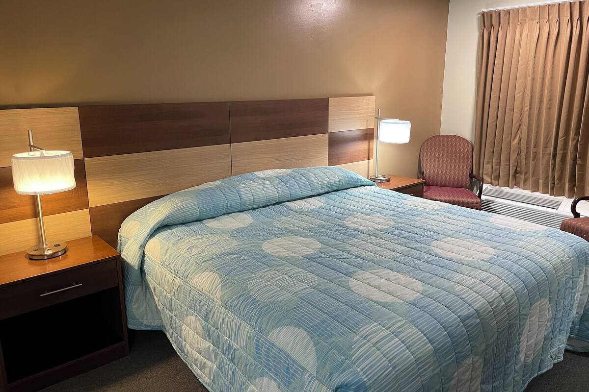 Habitación de hotel con una cama grande, una mesita de noche con lámpara, una silla y un ventanal con cortinas.