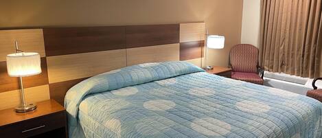 Quarto de hotel com uma cama grande, mesa de cabeceira com abajur, uma cadeira e uma janela com cortinas.