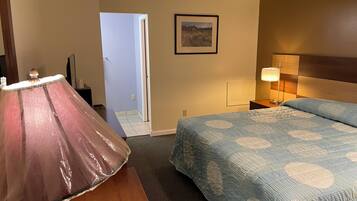 A hotel room with a bed, a nightstand with a lamp, a framed picture on the wall, and a door leading to another room.
