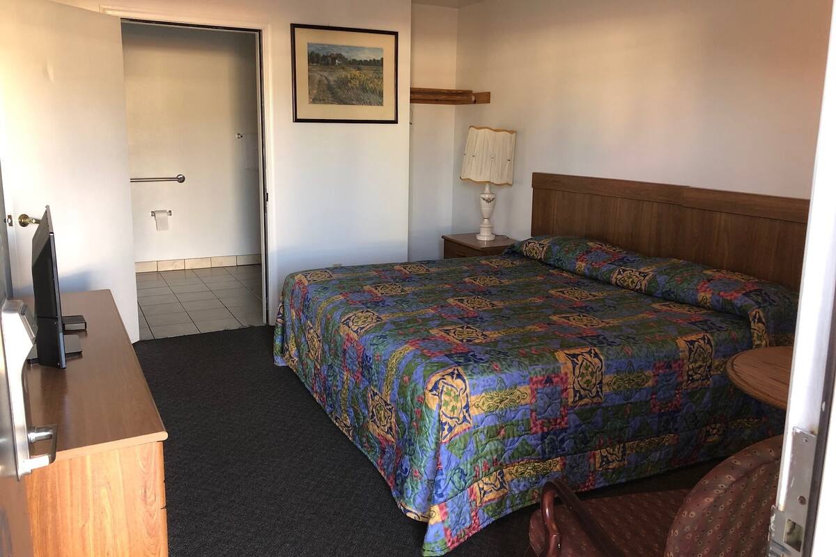Una habitación de hotel con cama, escritorio, silla, lámpara y puerta que da a un baño.