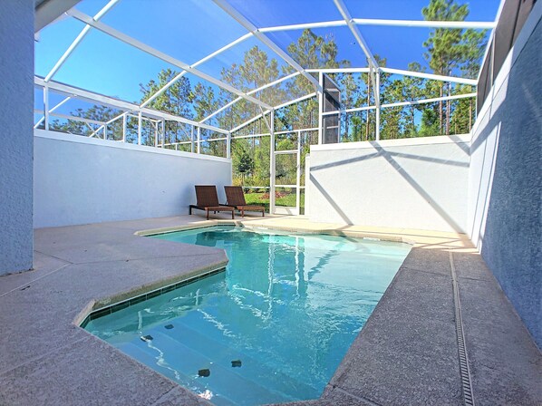A swimming pool with a glass enclosure, two lounge chairs, and a view of trees.