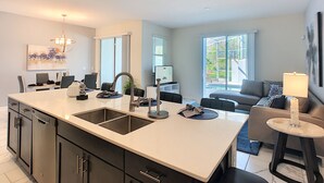 A modern kitchen with a large island, a dining area with chairs, and a living room with a sofa and a lamp. - Ocean Breeze by Florida Spirit (Kissimmee)