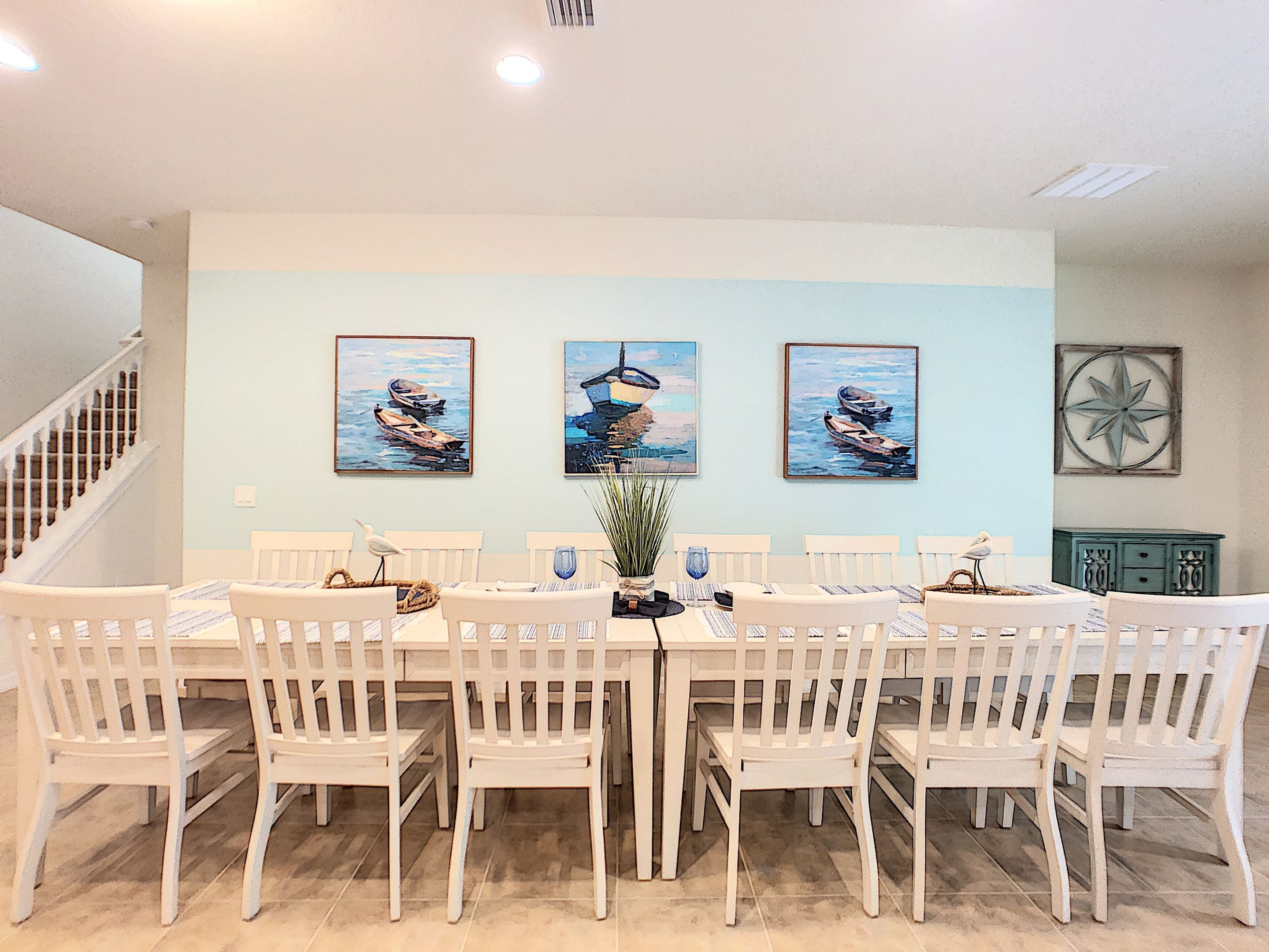 Un espace repas avec une longue table, des chaises blanches et trois tableaux accrochés au mur.