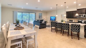 A modern kitchen with a central island, dining area with a large table and chairs, and a living area with a sofa and television.
