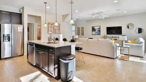 A modern kitchen with stainless steel appliances, a central island, pendant lighting, and a connected living area with a sofa and TV. - Lotus Garden by Florida Spirit (Kissimmee)