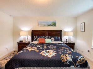 A bedroom with a large bed, two bedside tables, a nightstand, a painting above the bed, and a window on the right.