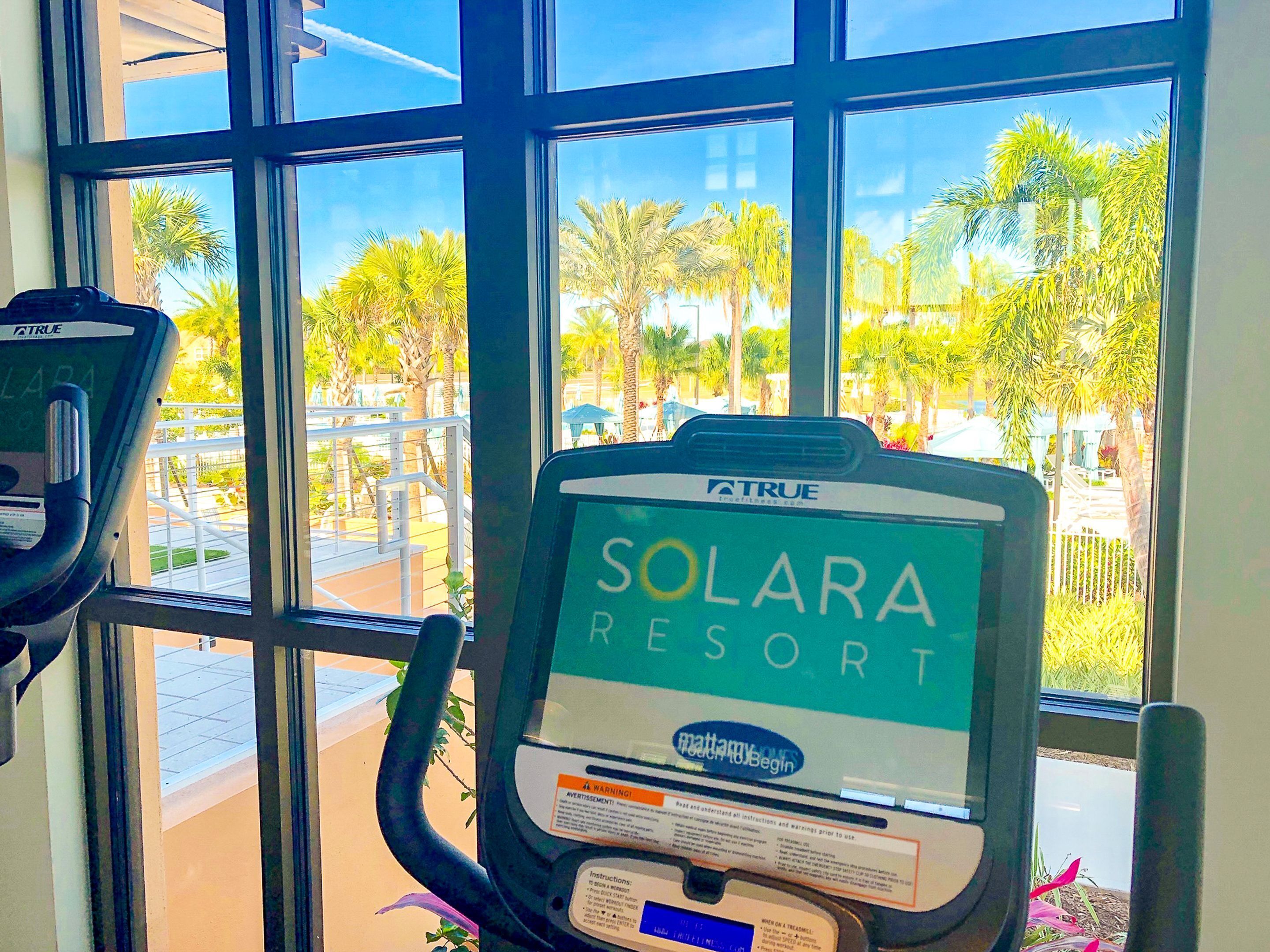 Ein Laufband mit einem Bildschirm, der die Logos „SOLARA RESORT“ und „Mattamy Homes“ zeigt, steht in der Nähe eines Fensters mit Blick auf Palmen und einen Poolbereich.
