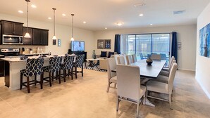 A modern kitchen with dark cabinets, a central island with bar stools, a dining area with a large table and chairs, and a TV mounted on the wall. - Bengal Sunrise by Florida Spirit (Kissimmee)