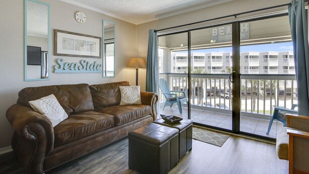 A living room with a brown leather sofa, a small black ottoman, and a blue chair. There is a large sliding glass door leading to a balcony with a view of a building labeled "BEACH". - Casa Del Mar Beach Suites (Galveston)