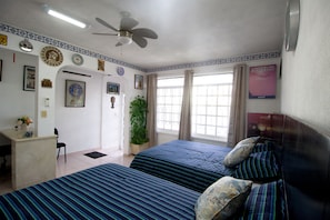 Quarto de hotel com duas camas, uma escrivaninha, uma cadeira, ventilador de teto, planta em vaso e janela com cortinas.