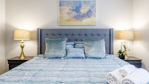 Ein ordentlich bezogenes Bett mit Streifenmuster, zwei Kissen und einem Kopfteil. Es gibt zwei Nachttische, auf denen Lampen und eine kleine Pflanze stehen.