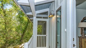 Una terraza de madera con barandal blanco, un cristal que da a una habitación y una lámpara sobre un mueble de madera.