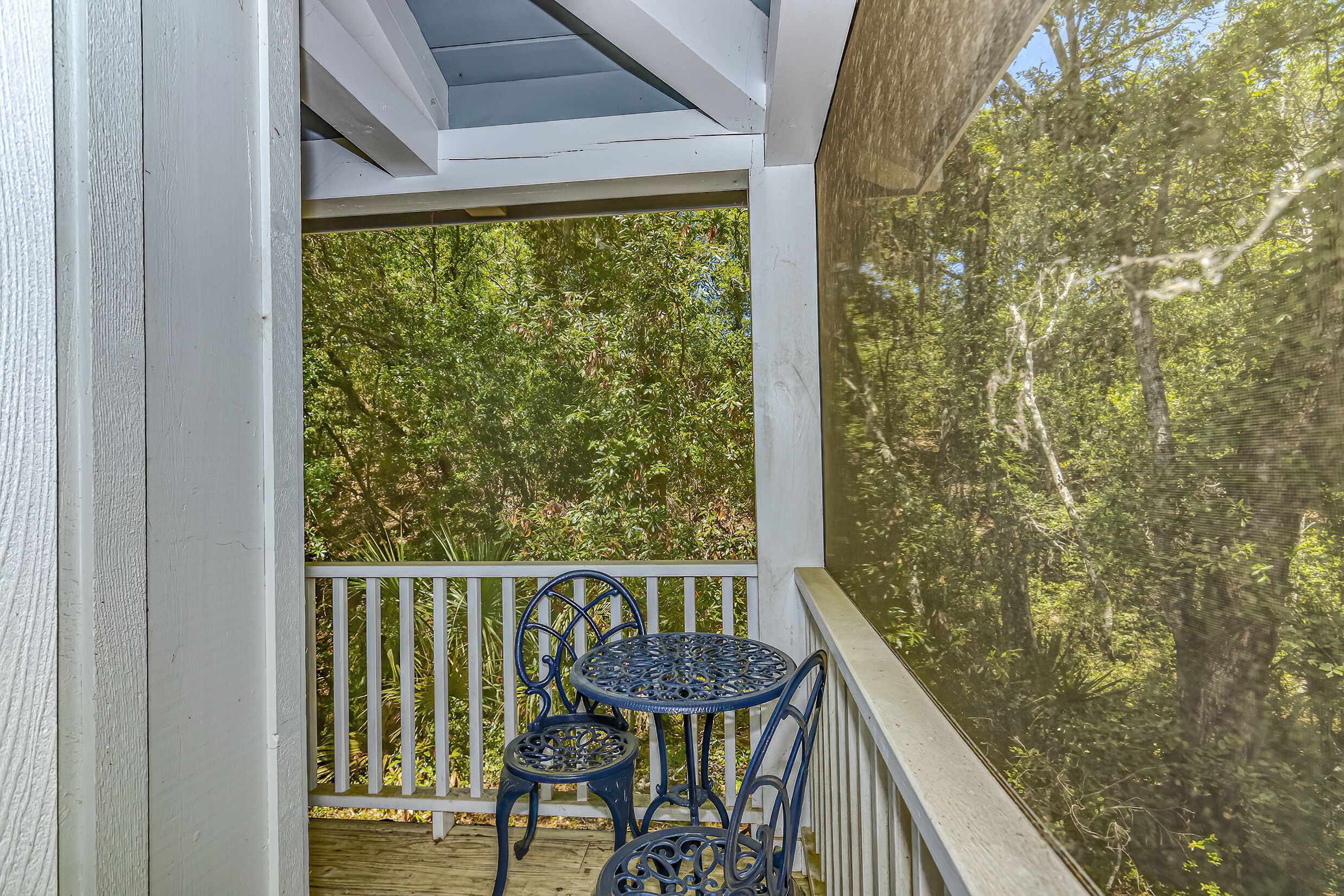 A covered outdoor area with a table and chairs, surrounded by a white railing and a view of dense trees.