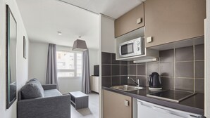 Cocina compacta con microondas, pileta y tetera. Hay un sofá gris y una pequeña mesa en la zona de estar.