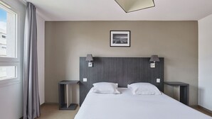 Una habitación de hotel con una cama grande, dos mesitas de noche, un cabecero con un cuadro y una ventana con cortinas.