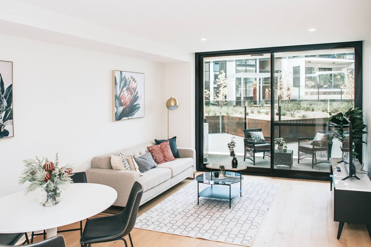 Ein modernes Wohnzimmer mit Sofa, Couchtisch und Essbereich mit Stühlen und Tisch. Ein großes Fenster bietet Blick auf Gartenmöbel und Pflanzen.
