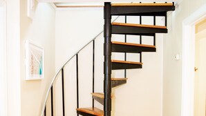 Una escalera de caracol con escalones de madera y barandillas de metal negro en una habitación con paredes de tonos claros y suelo de madera.
