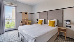 Hotellihuoneessa on suuri sänky, työpöytä ja tuoli. Huoneessa on ikkuna, josta avautuu näkymä ulkona oleviin rakennuksiin.