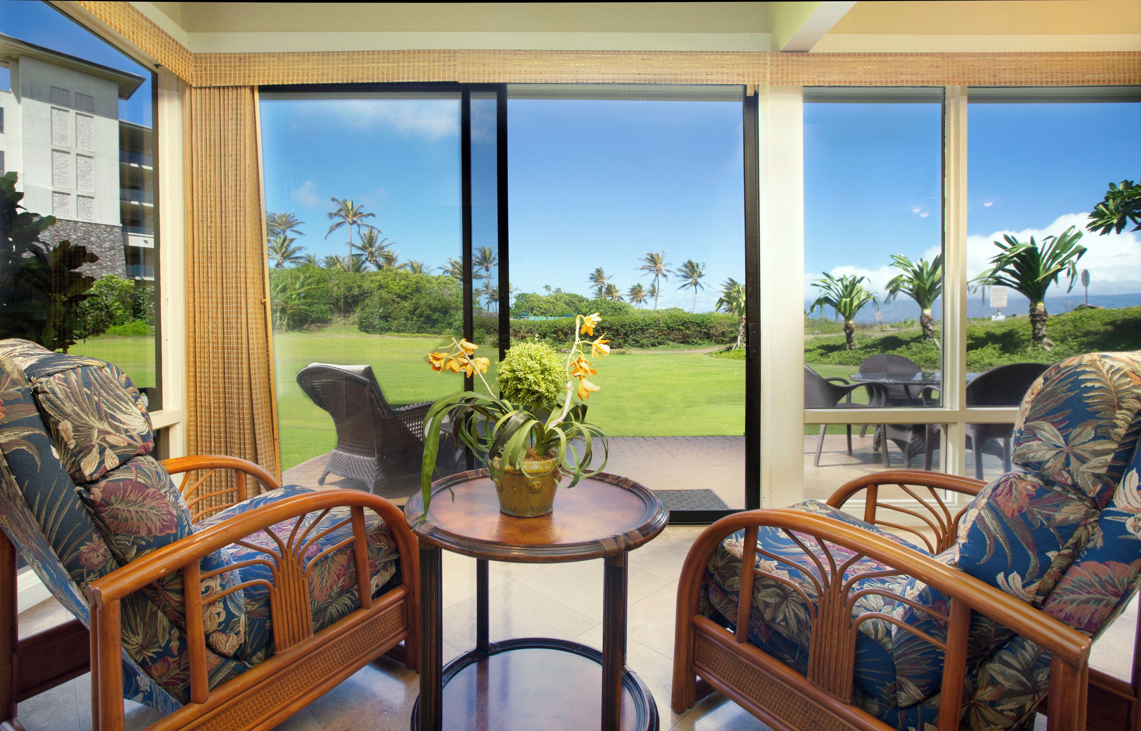 Una habitación con muebles de mimbre, una mesita con planta en maceta y ventanas amplias que dan a un paisaje verde con palmeras.