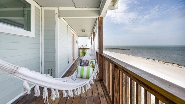 A wooden deck with a hammock, a small seating area with pillows, and a view of the beach and ocean. - SUNRISE ON MAG - Beach House (Port Lavaca)