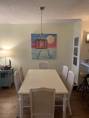 A dining area with a white table and wicker chairs, a hanging lantern, and a wall-mounted painting.