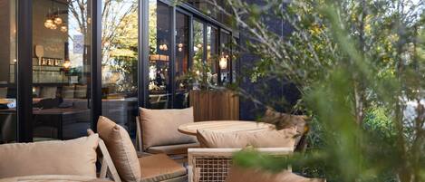 Un área de estar al aire libre con mobiliario de madera y cojines, ventanas amplias y luz interior visible a través del vidrio.