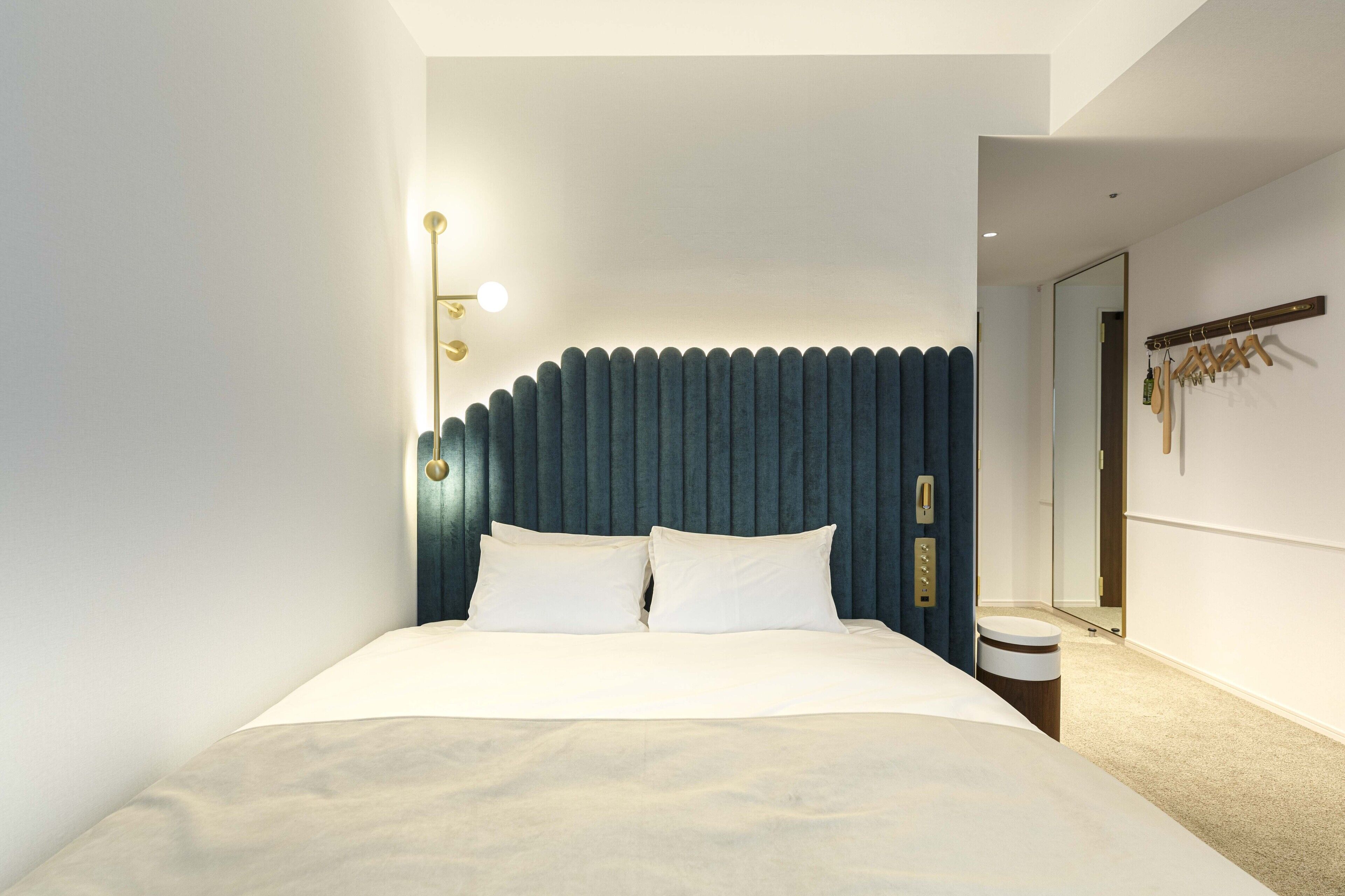 A modern hotel room with a large bed, a headboard with vertical lines, a bedside table, a mirror, and a coat rack.