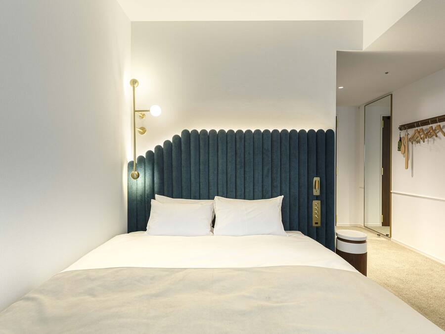 A modern hotel room with a large bed, a headboard with vertical lines, a bedside table, a mirror, and a coat rack.